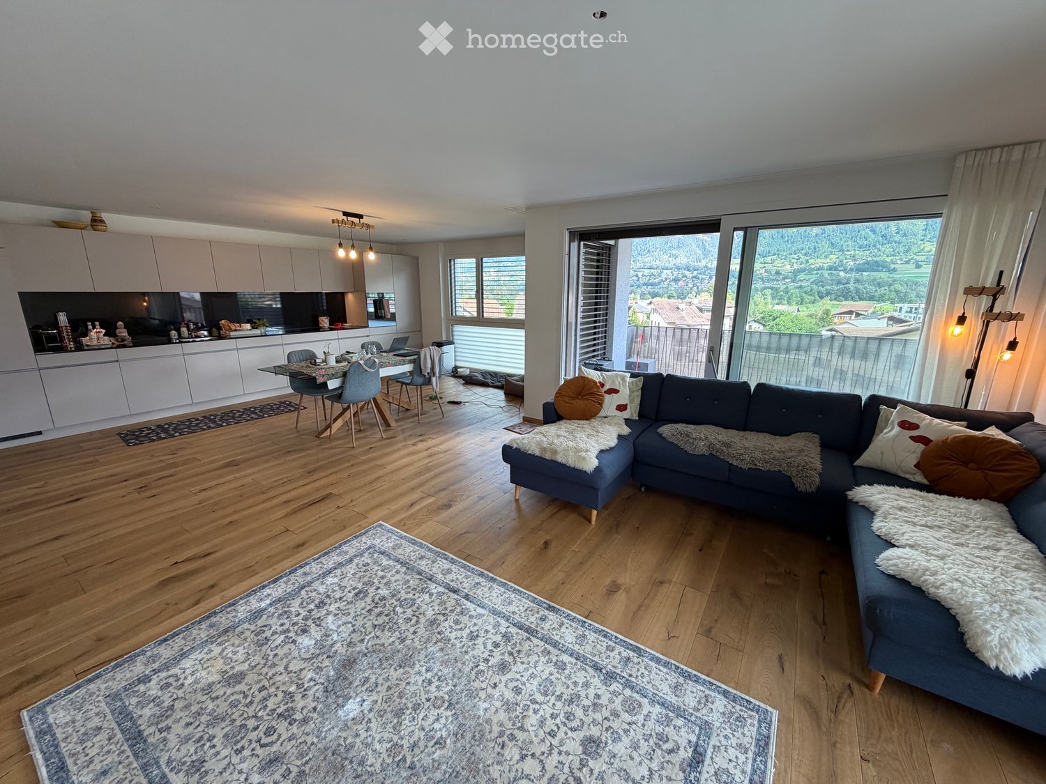 Modern open-plan living and dining area with wooden flooring, large windows offering a scenic view, a blue sectional sofa, a dining table with chairs, and a modern kitchen with white cabinets and stainless steel appliances.