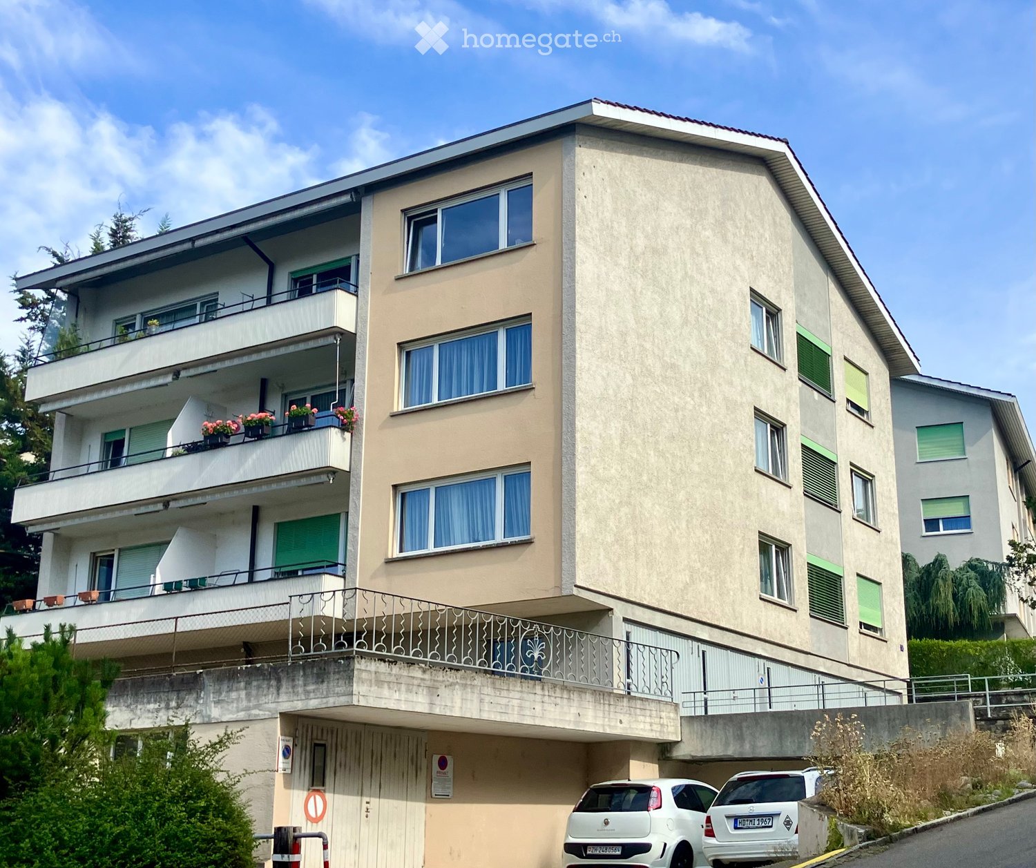 Four-story residential building with balconies, large windows, and a garage. Beige facade with green shutters and flower pots on balconies.