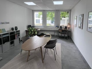 Modern office or meeting room with a large wooden table, chairs, shelves, a small plant, and a lamp. The space features large windows and a clean, minimalist design.