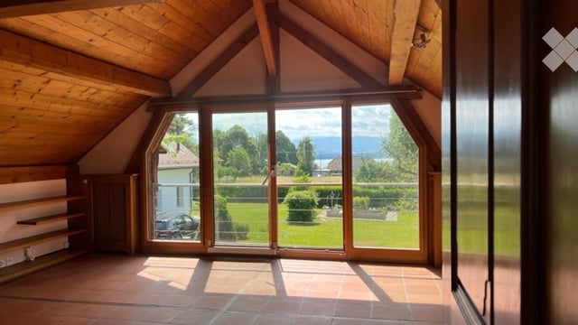 Bright, airy attic space with wooden beams, large glass doors opening to a green garden view, tiled floor, and built-in wooden shelving unit.