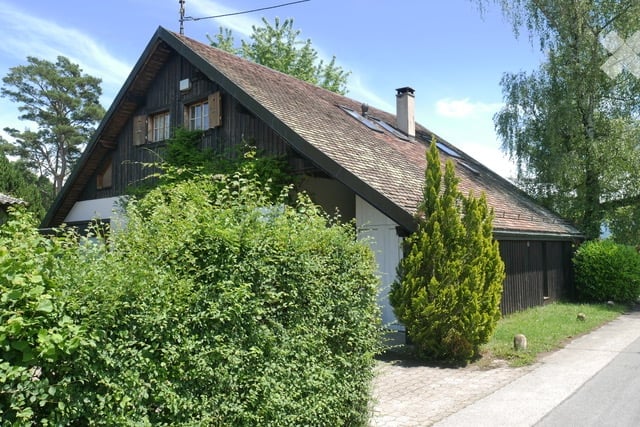 Traditional wooden house with a steeply pitched roof, surrounded by greenery and bushes.