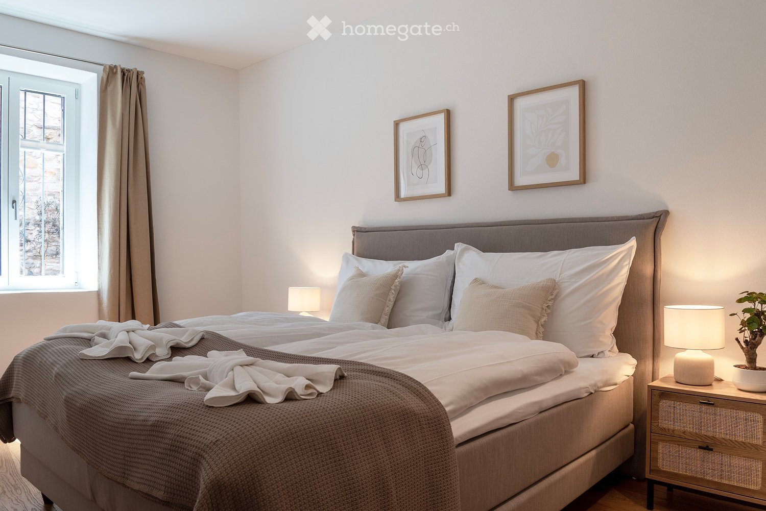 Spacious bedroom with large window, gray upholstered bed frame, white bedding, gray throw blanket, two framed artwork on the wall, and a bedside table with lamps