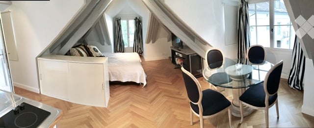 Cozy attic room with wooden herringbone flooring, a bed, a small dresser, a glass dining table with chairs, and a television on a stand.