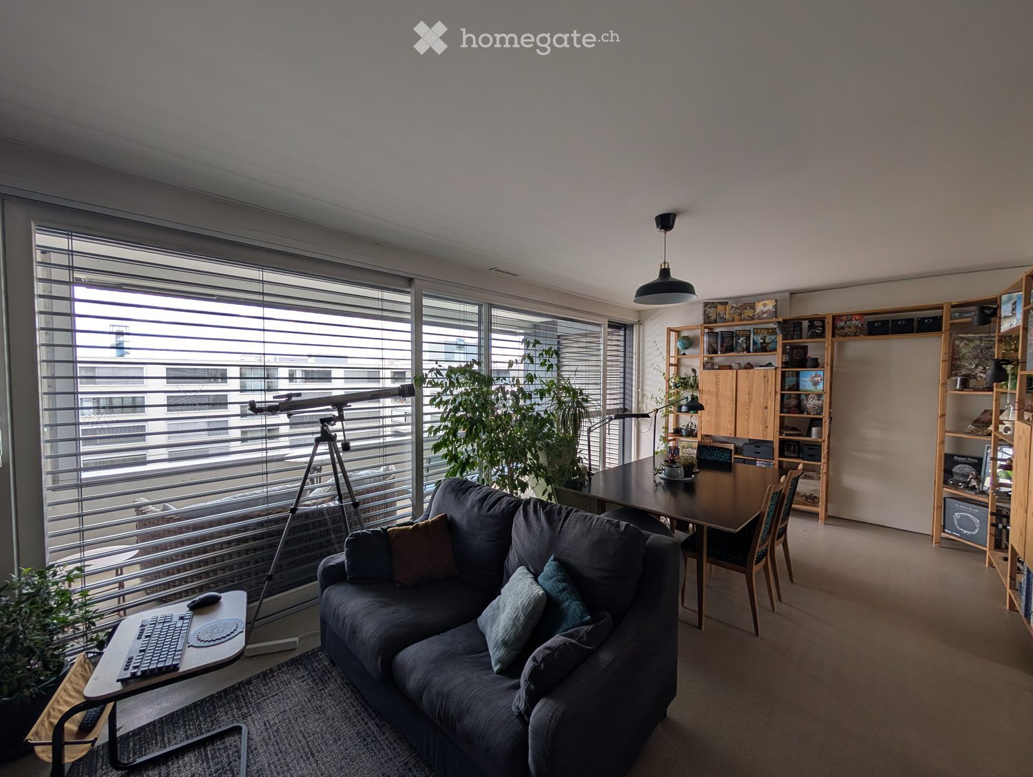 Modern living room with large windows, grey sofa, laptop on side table, dining table with chairs, bookshelf, and potted plants.