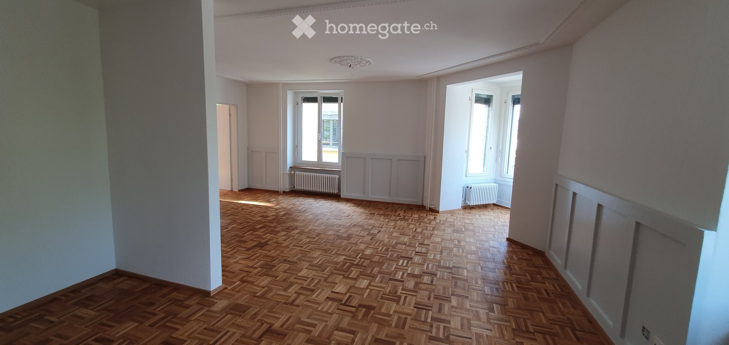 Empty room with wooden herringbone flooring, white walls, and large windows allowing natural light. Radiators are visible under the windows.