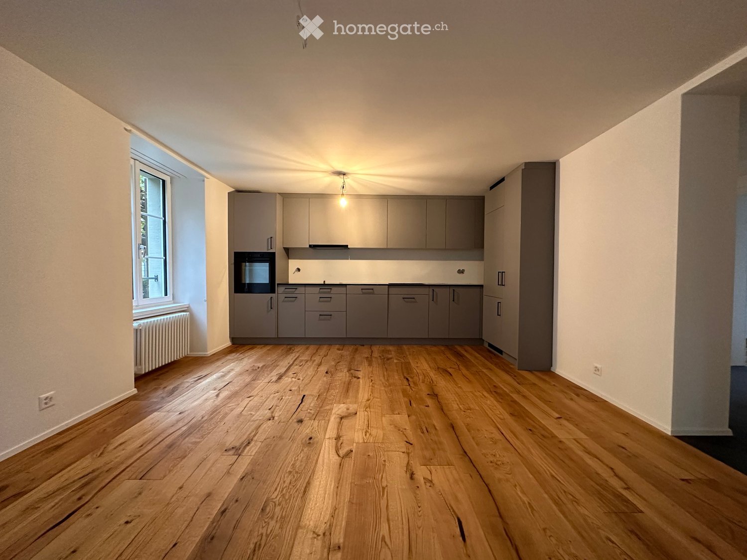 Empty kitchen with light grey cabinets, wooden flooring, and a window on the left side.