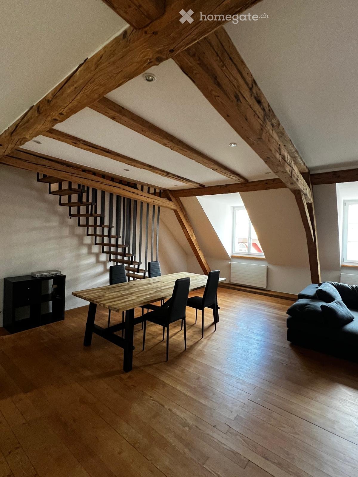 Open-plan living and dining area with exposed wooden beams, hardwood flooring, a sofa, a dining table with chairs, and a cabinet.