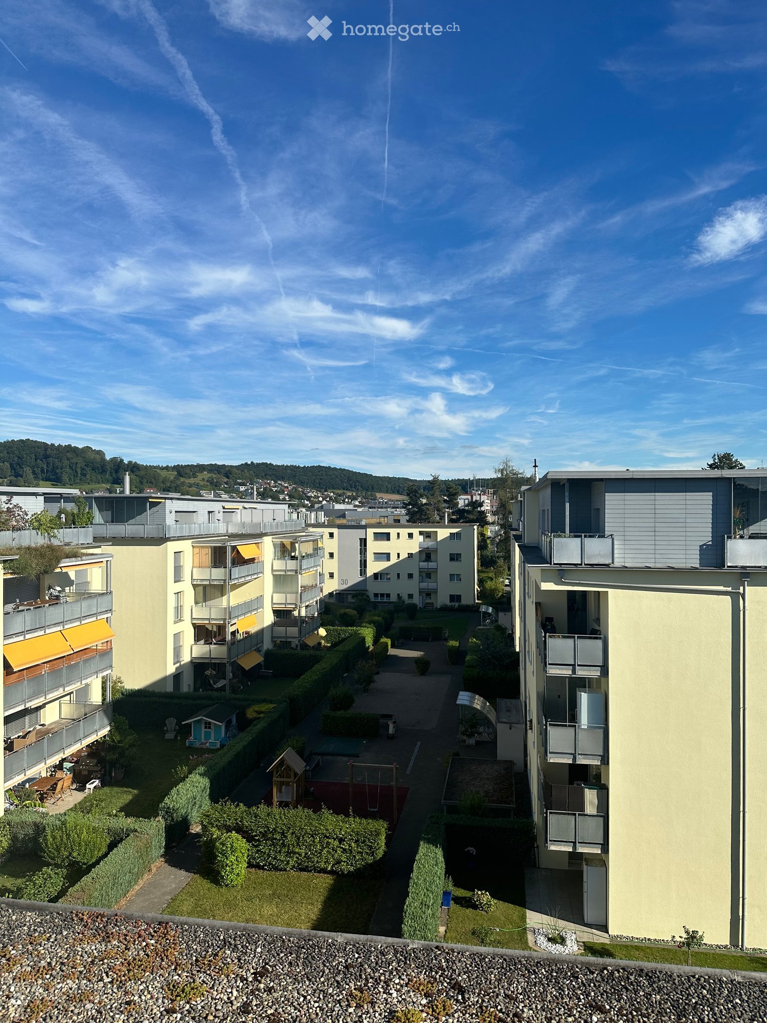 Courtyard view of a residential complex with multiple apartment buildings, balconies, greenery, and outdoor seating areas.