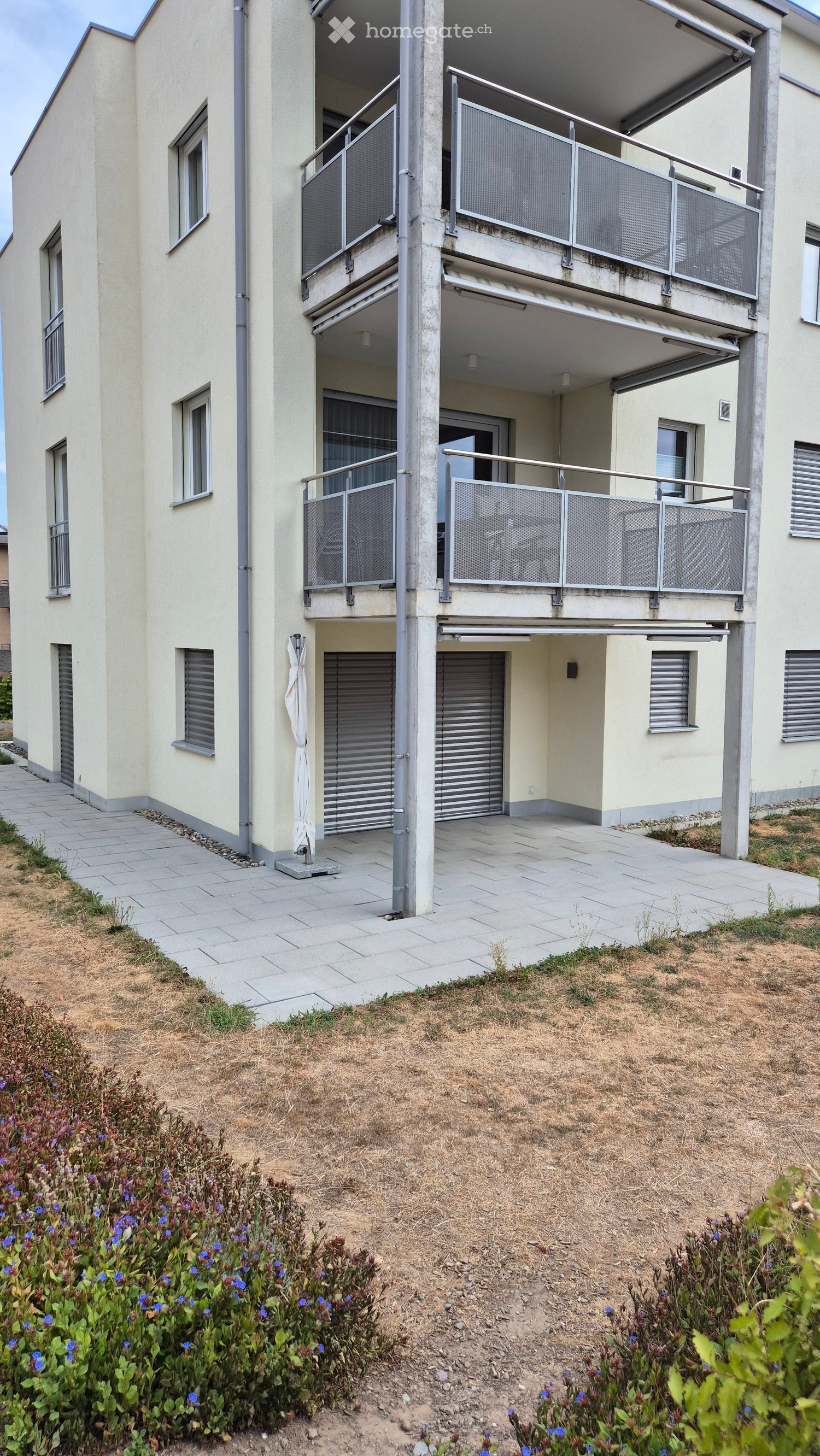 Modern multi-story building with balconies, grey metal railings, and a paved walkway.
