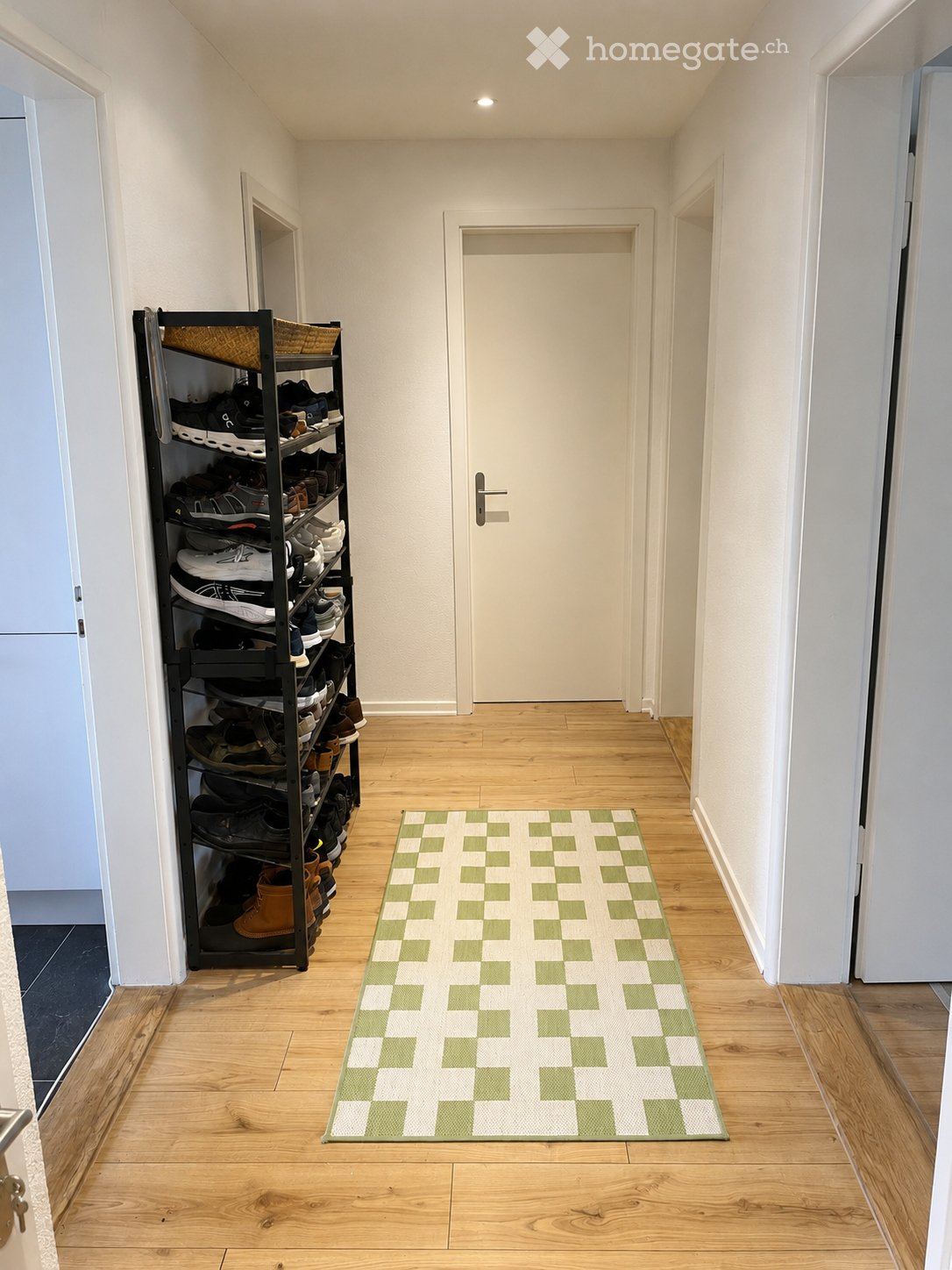 A narrow hallway with wooden flooring, white walls, and a green and white checkered rug. A black shoe rack filled with various shoes is positioned on the left side, and a closed white door is visible at the end of the hallway.