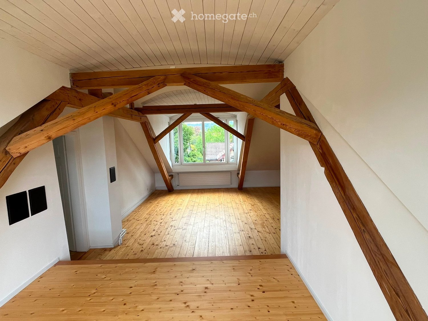 An empty attic space with wooden beams, wooden flooring, and large windows offering a view of greenery outside.