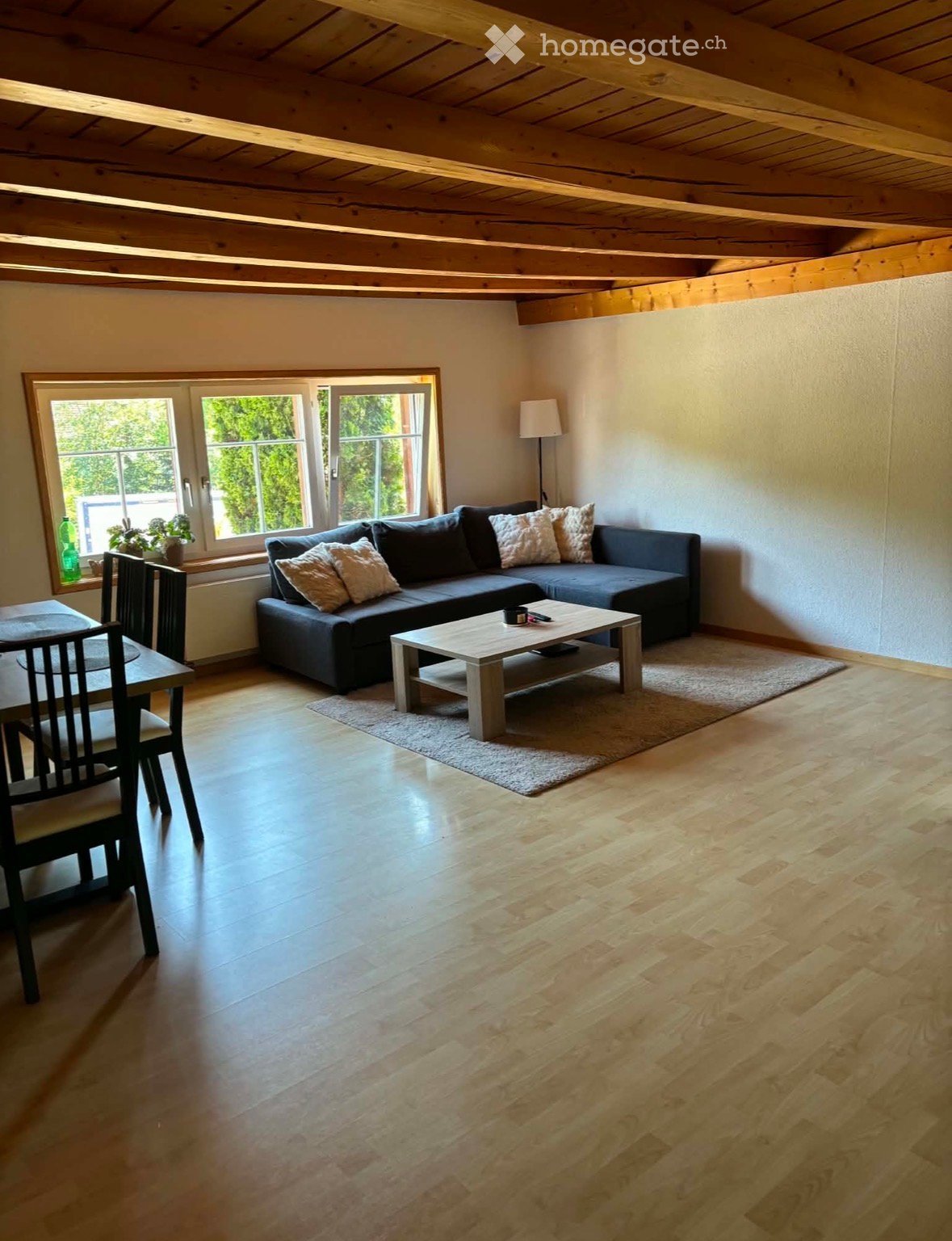 Bright living room with wooden floors, exposed wooden ceiling beams, large windows, grey sectional sofa, coffee table, dining area with table and chairs, and a floor lamp.