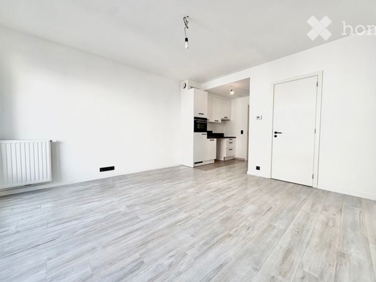 Empty room with white walls, light wooden flooring, and a white radiator on one wall. A doorway leads to a small kitchen area with white cabinets and a black oven.