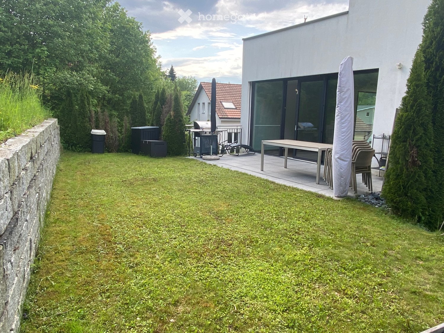 Well-maintained garden with a stone wall, lawn, patio area with a table and chairs, and a modern house with large glass doors.