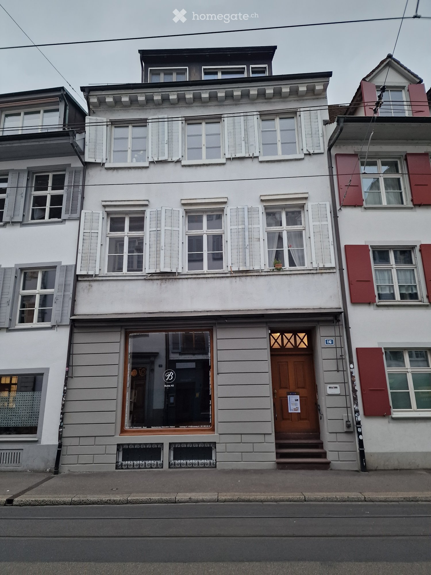 Three-story residential building with white facade, multiple windows with shutters, and a small balcony on the second floor. The ground floor features a storefront with glass windows and a wooden entrance door.