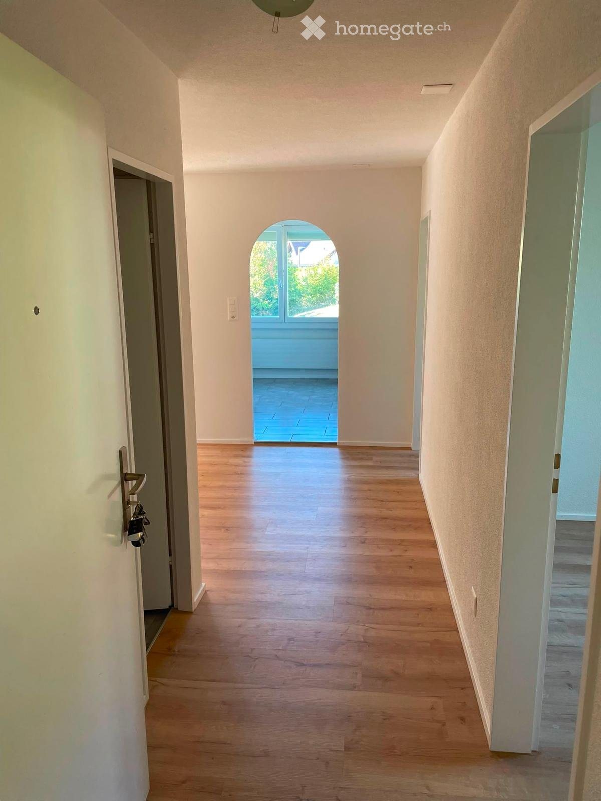 Empty hallway with wooden flooring, white walls, and a small arched window at the end leading to a blue-tiled area.