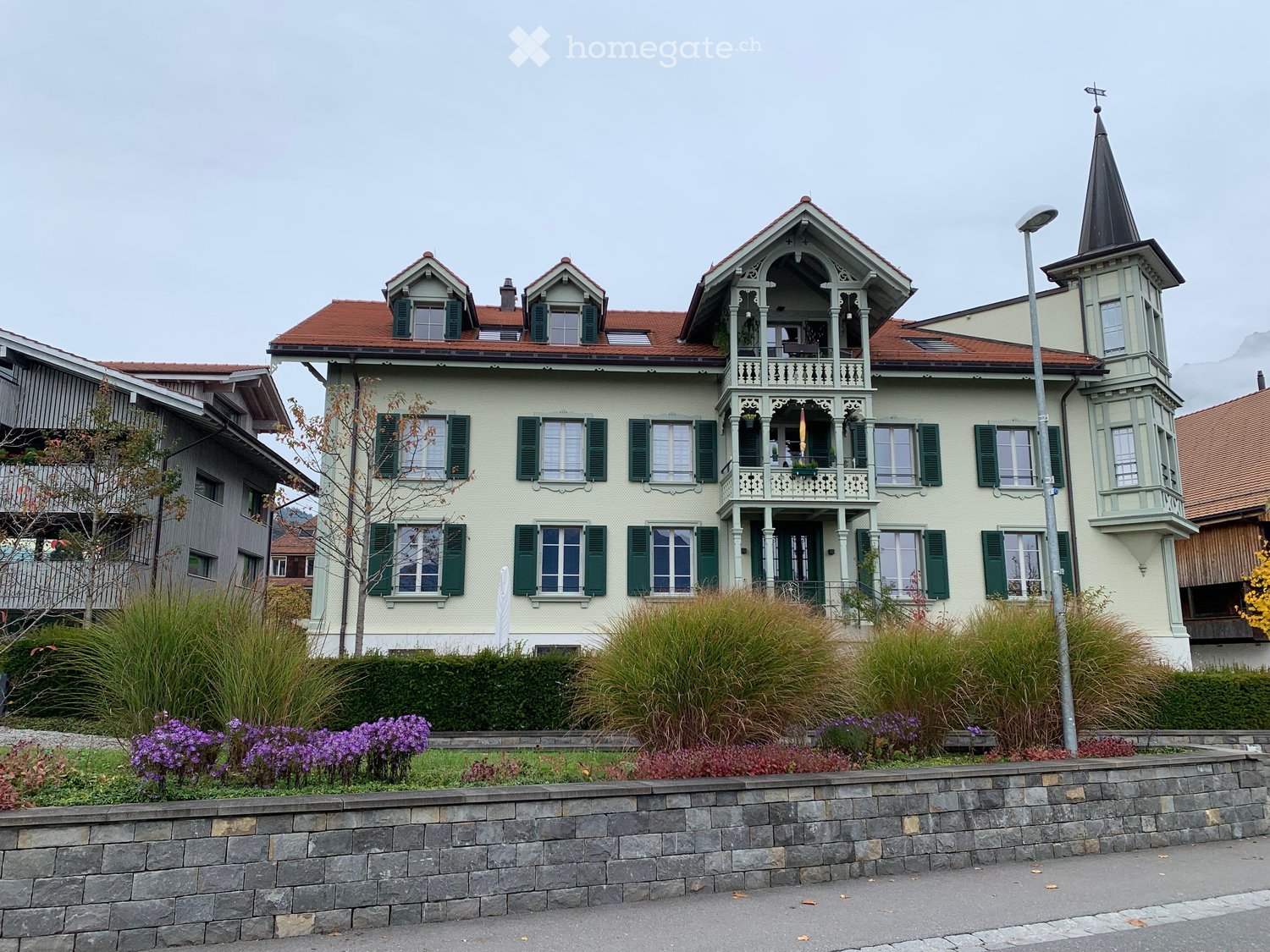A two-story house with a light yellow facade, green shutters, and a red-tiled roof. Features include a balcony with ornate railings, a tower with a pointed roof, and a well-maintained garden with purple flowers and tall grasses.
