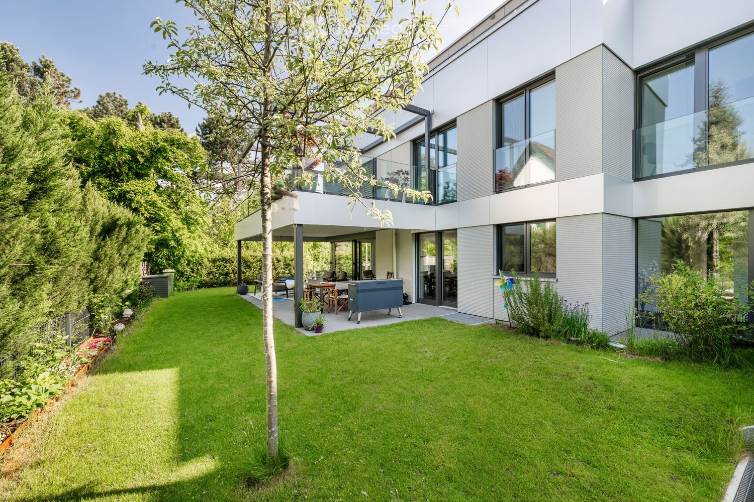 Modern house with a spacious garden, featuring a well-maintained lawn, patio area with outdoor furniture, and a barbecue. The house has large windows and a sleek, contemporary design.