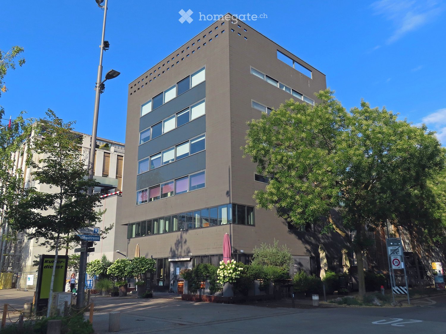 Modern multi-story building with large windows and a flat roof, surrounded by trees and greenery.