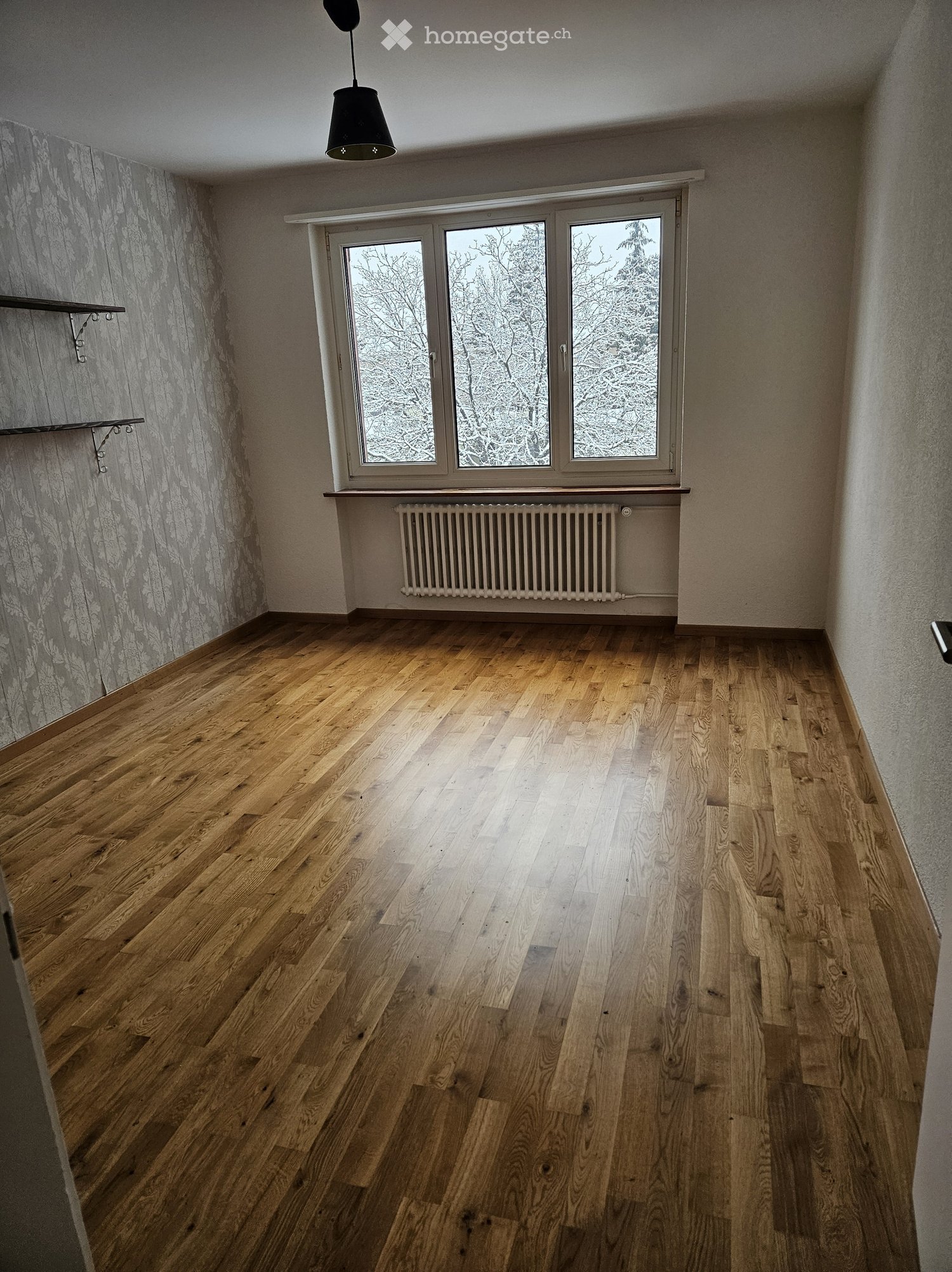 Empty room with wooden flooring, large window with frosted glass, radiator, and a hanging light fixture.