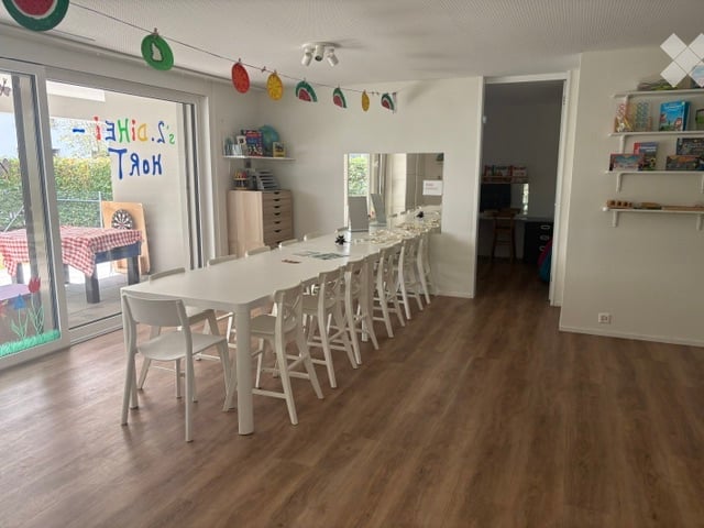 Bright room with wooden flooring, large white table surrounded by chairs, shelves with books and decorations, and a large window with a view of the garden.