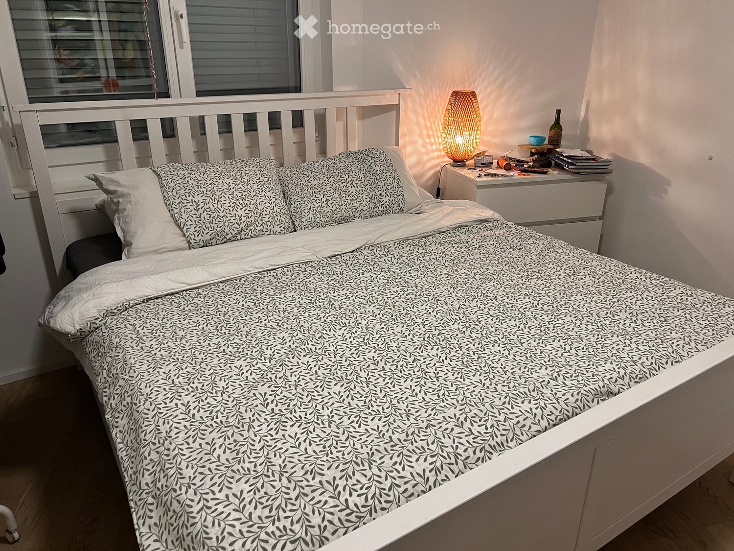 Cozy bedroom with a white bed, patterned bedding, a nightstand with a lamp, and a window with blinds.