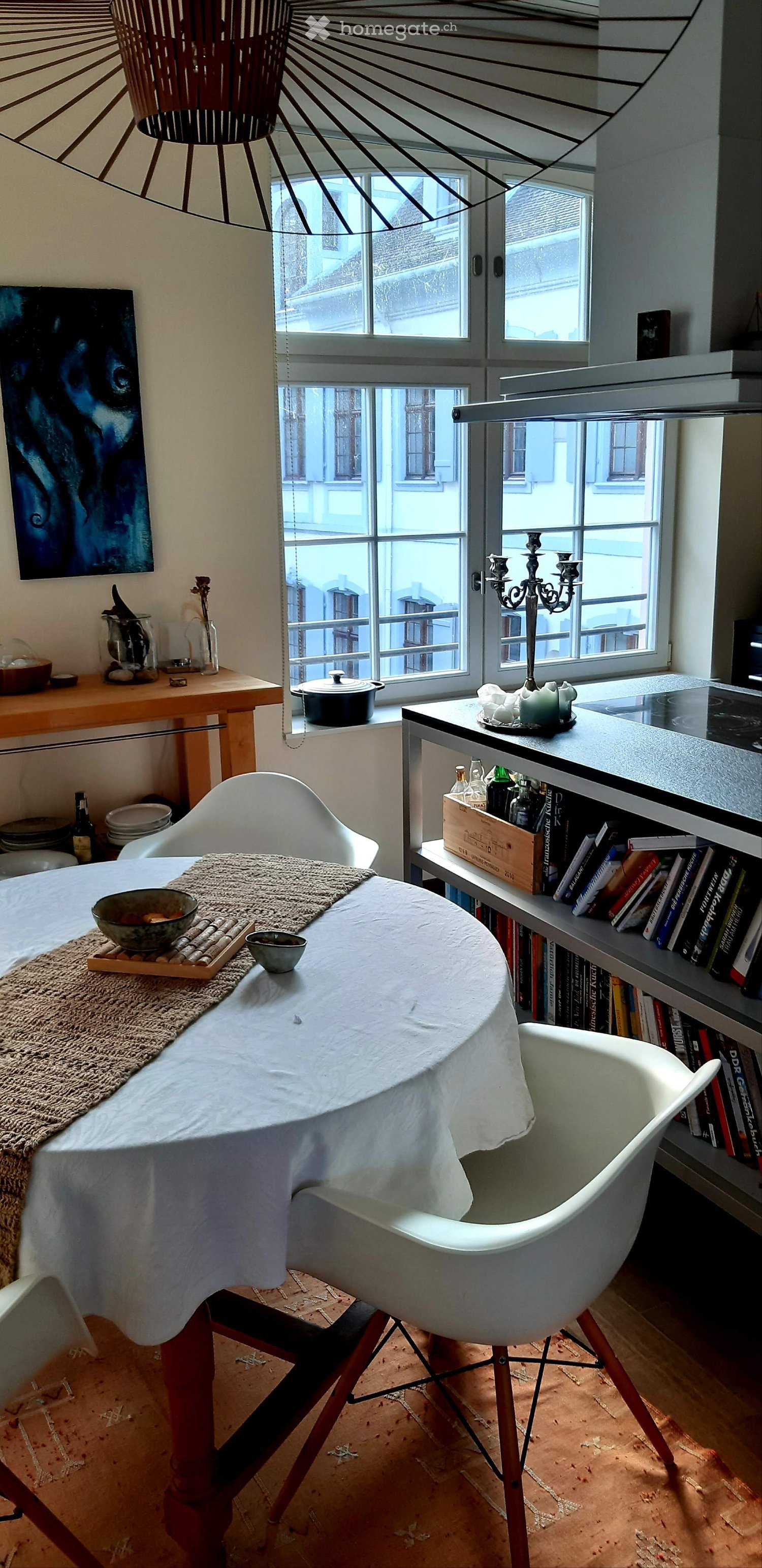 A modern, well-lit room with a dining table, chairs, bookshelf, and various decorative items. The room features large windows with a view of the street and a cozy, inviting atmosphere.