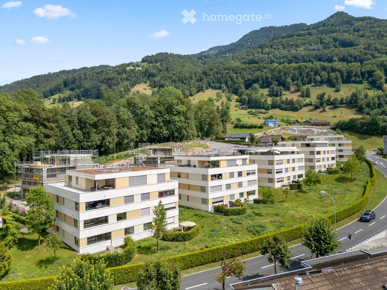 Aerial view of a modern residential complex with multiple buildings, surrounded by greenery and hills in the background.