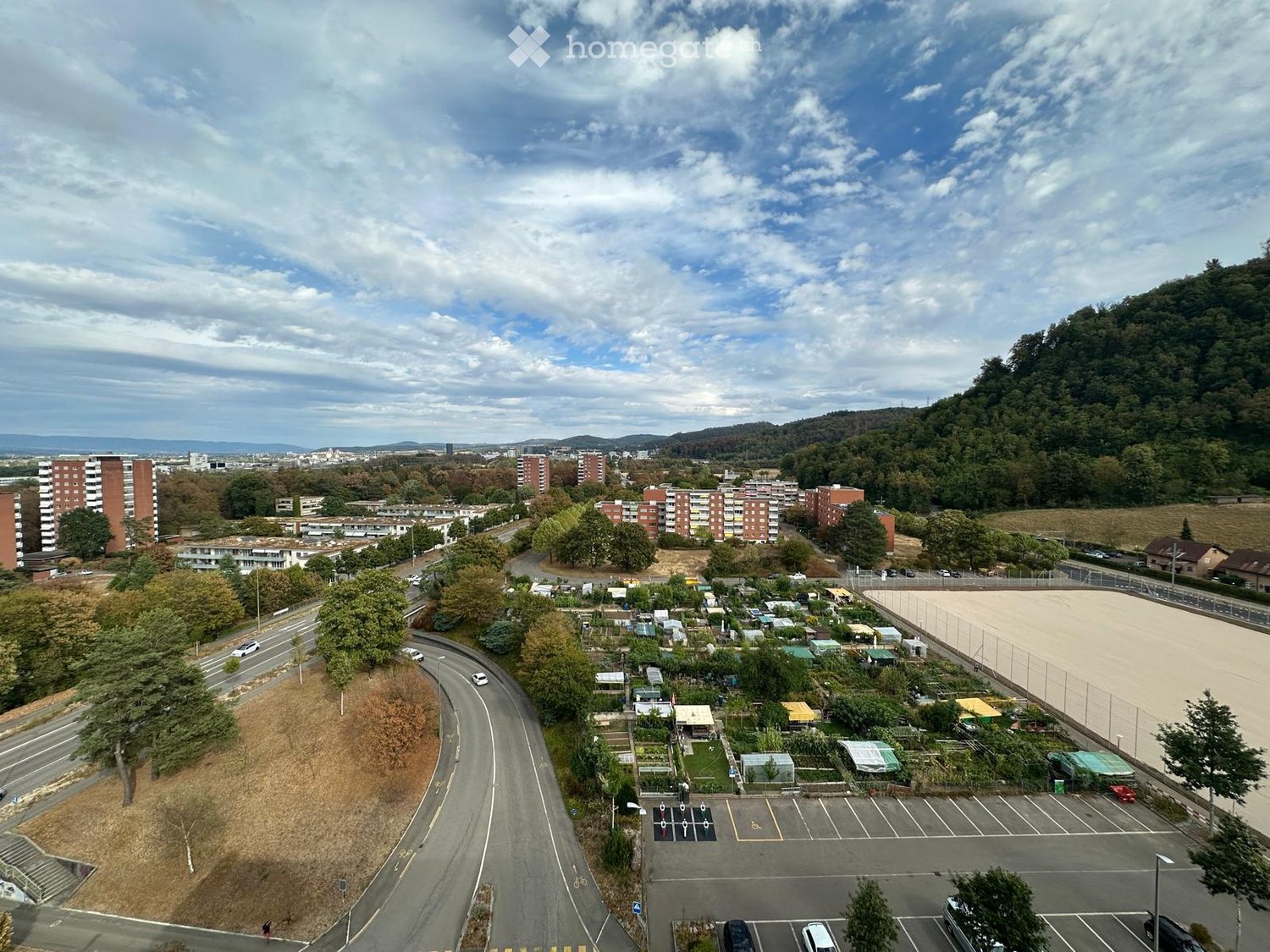 Aerial view of a residential area with apartment buildings, parking lots, a fenced sports field, and a garden area with small plots and greenhouses.