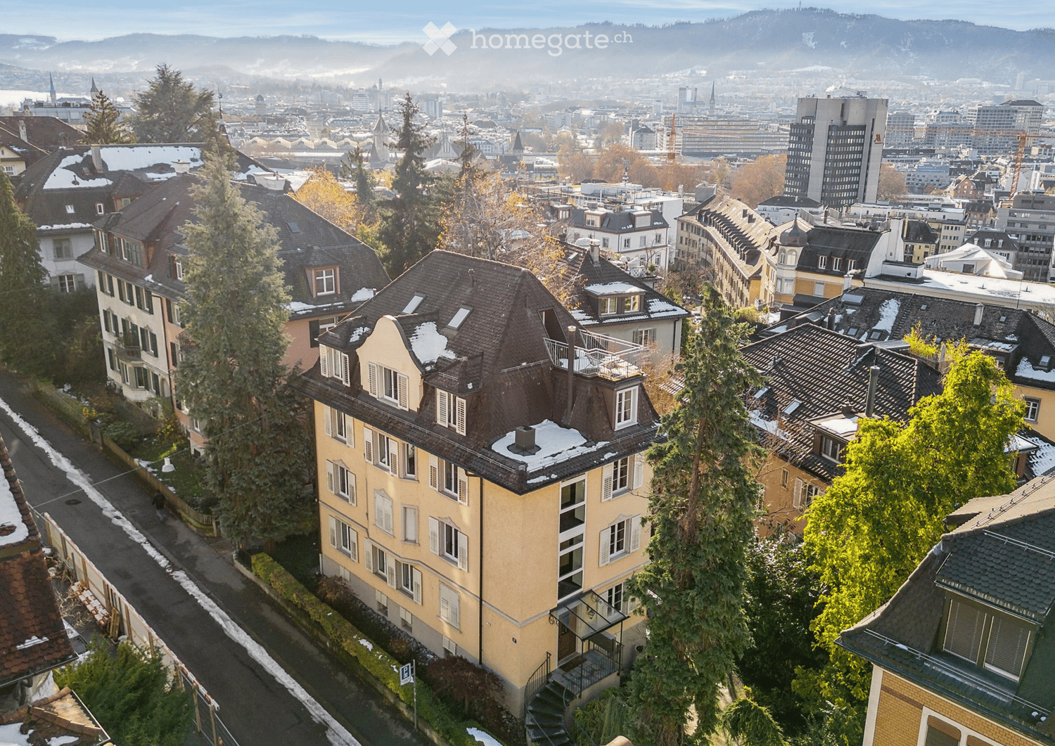 A multi-story residential building with a distinctive roof and white-framed windows, surrounded by other buildings and trees in a cityscape with distant mountains.
