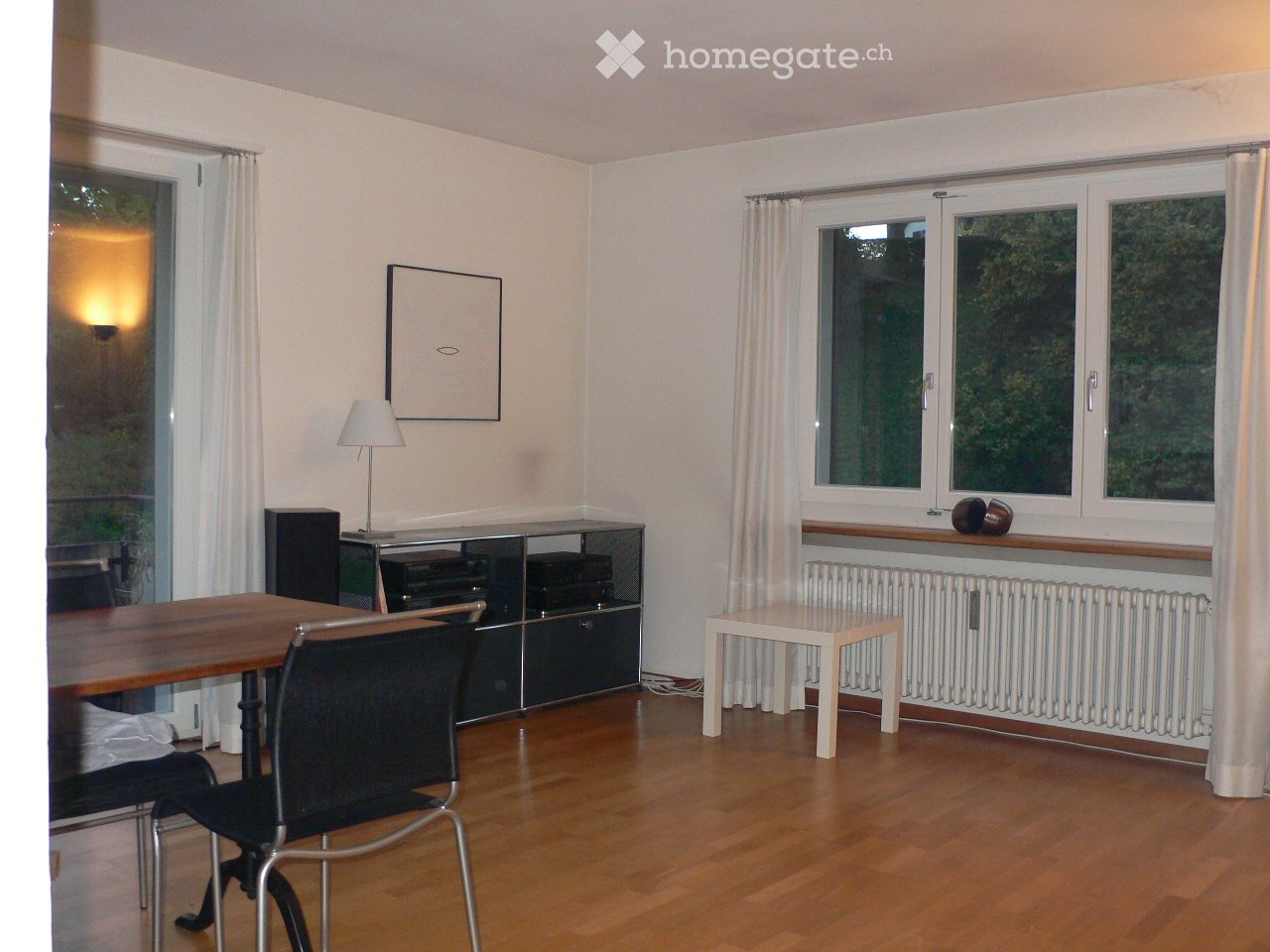 Bright room with wooden flooring, large windows with white curtains, a wooden dining table with chairs, a small white stool, a cabinet with electronic equipment, a lamp, and a picture frame on the wall.