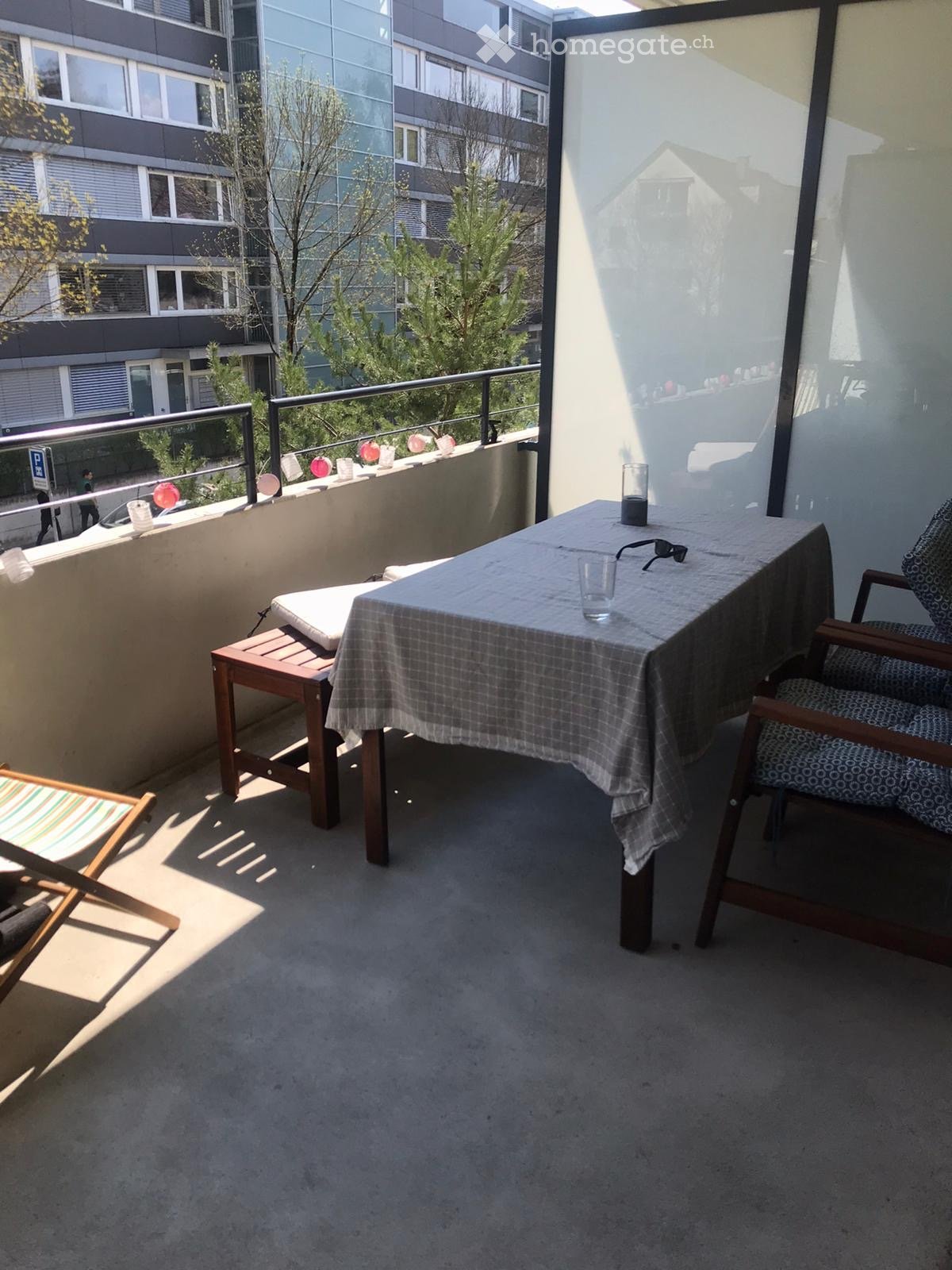 Balcony with a small table, two chairs, a stool, and a cup. The balcony has a metal railing and is decorated with colorful balloons.