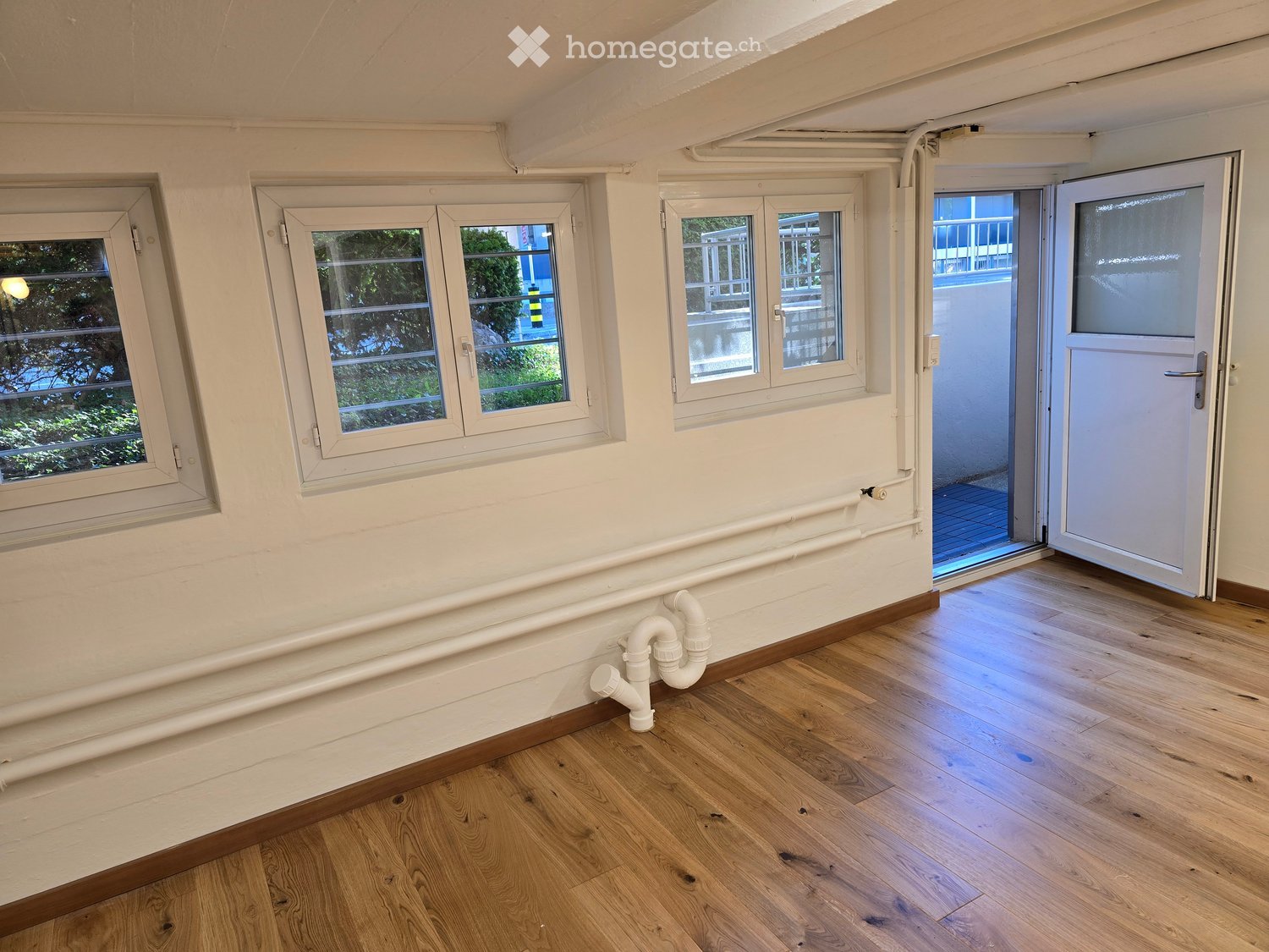 Empty room with wooden flooring, white walls, and multiple windows. Exposed white pipes run along the wall, and a door leads to an outdoor area.