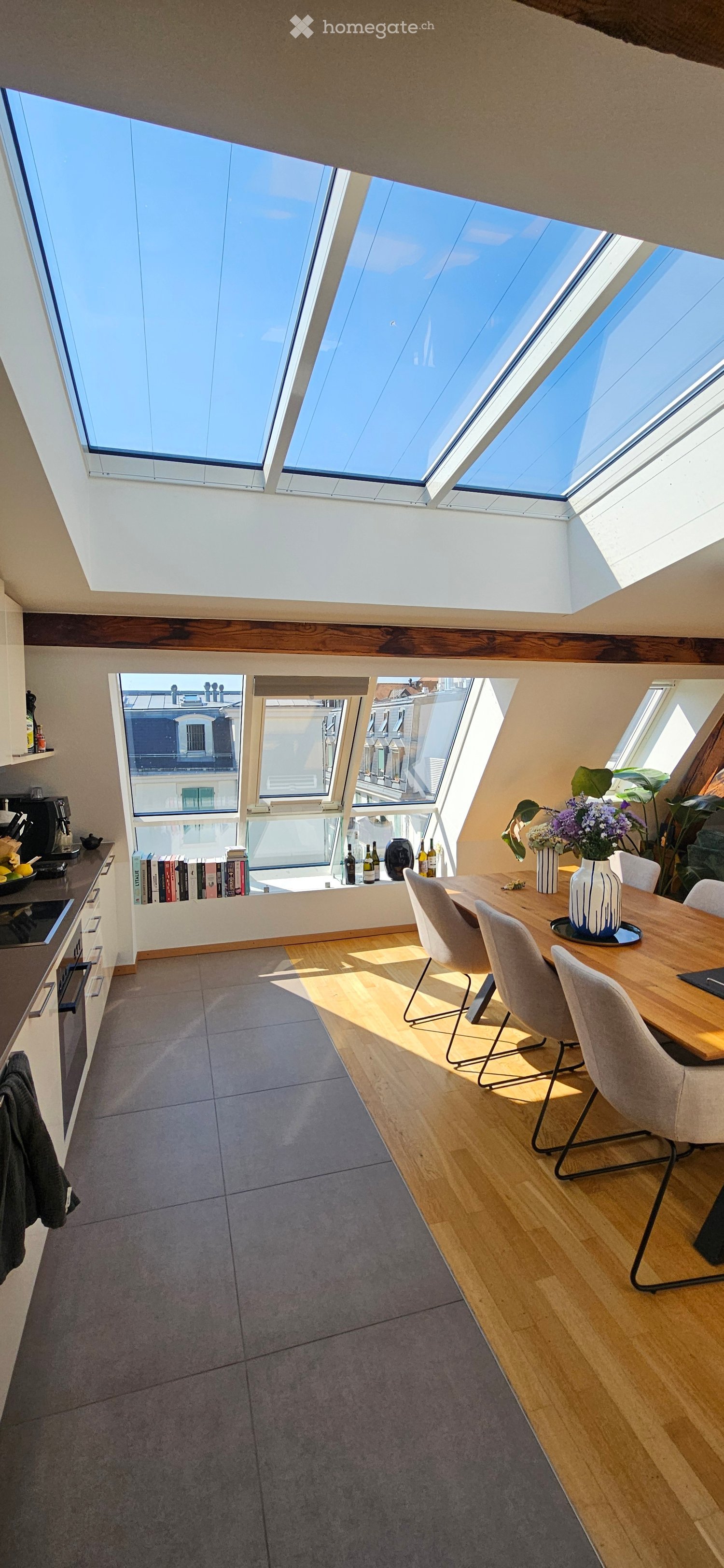 Bright, open-plan room with skylights, wooden dining table, modern chairs, kitchen area with appliances, and decorative vases.