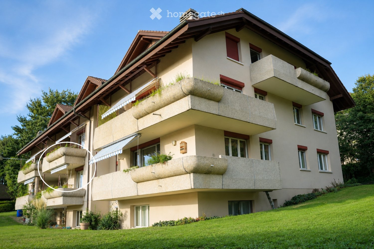 A two-story residential building with beige walls, multiple windows, and balconies with planters. The roof is brown with wooden beams, and the building is surrounded by a well-maintained lawn and trees.