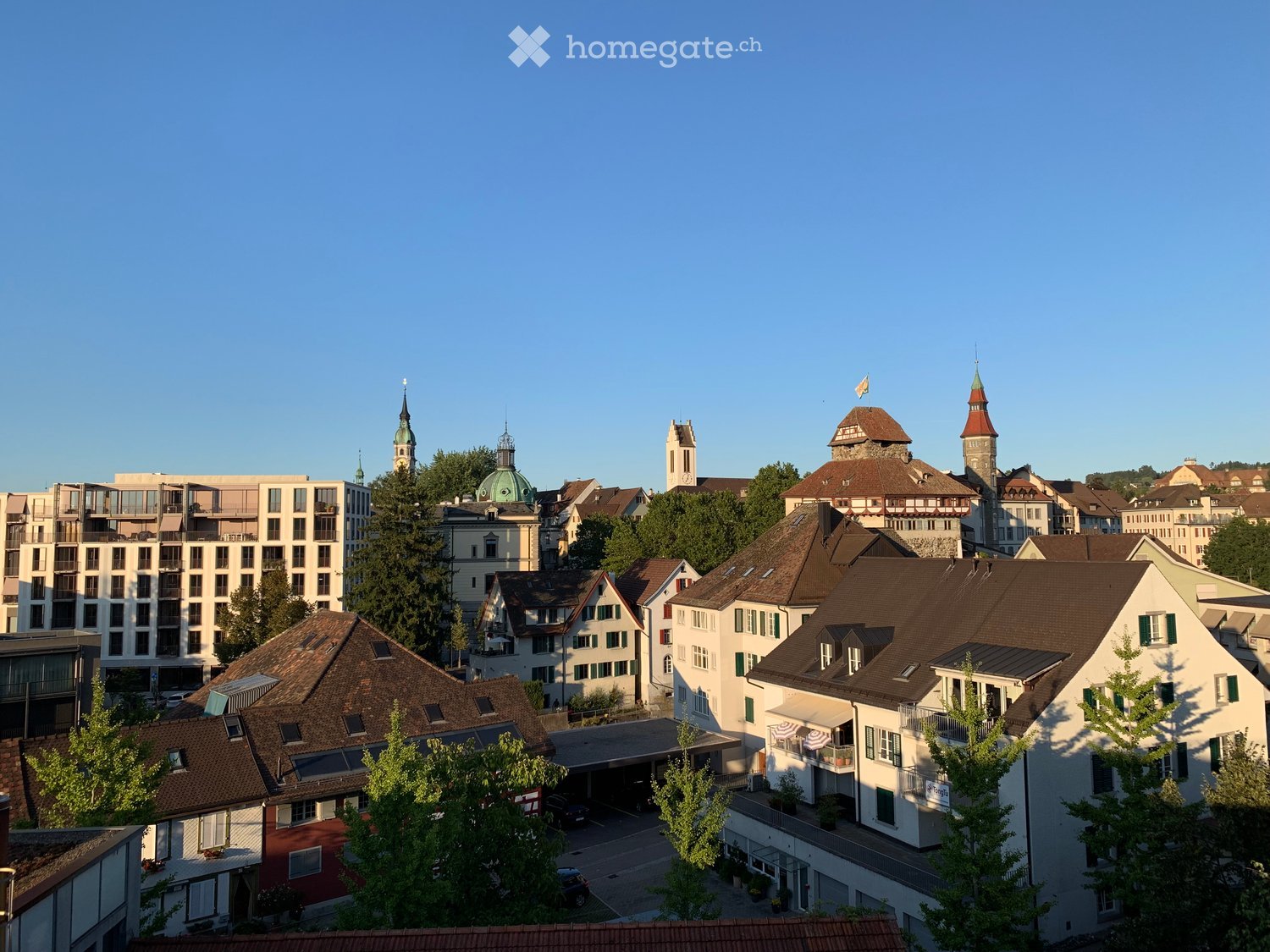 Cityscape with multiple buildings, some with spires, surrounded by trees, clear blue sky, a car parked in front of a building