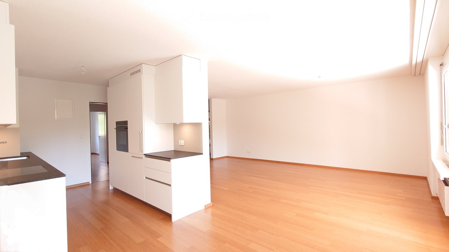 Empty room with white walls, wooden flooring, and a small kitchen area with white cabinets and a countertop.