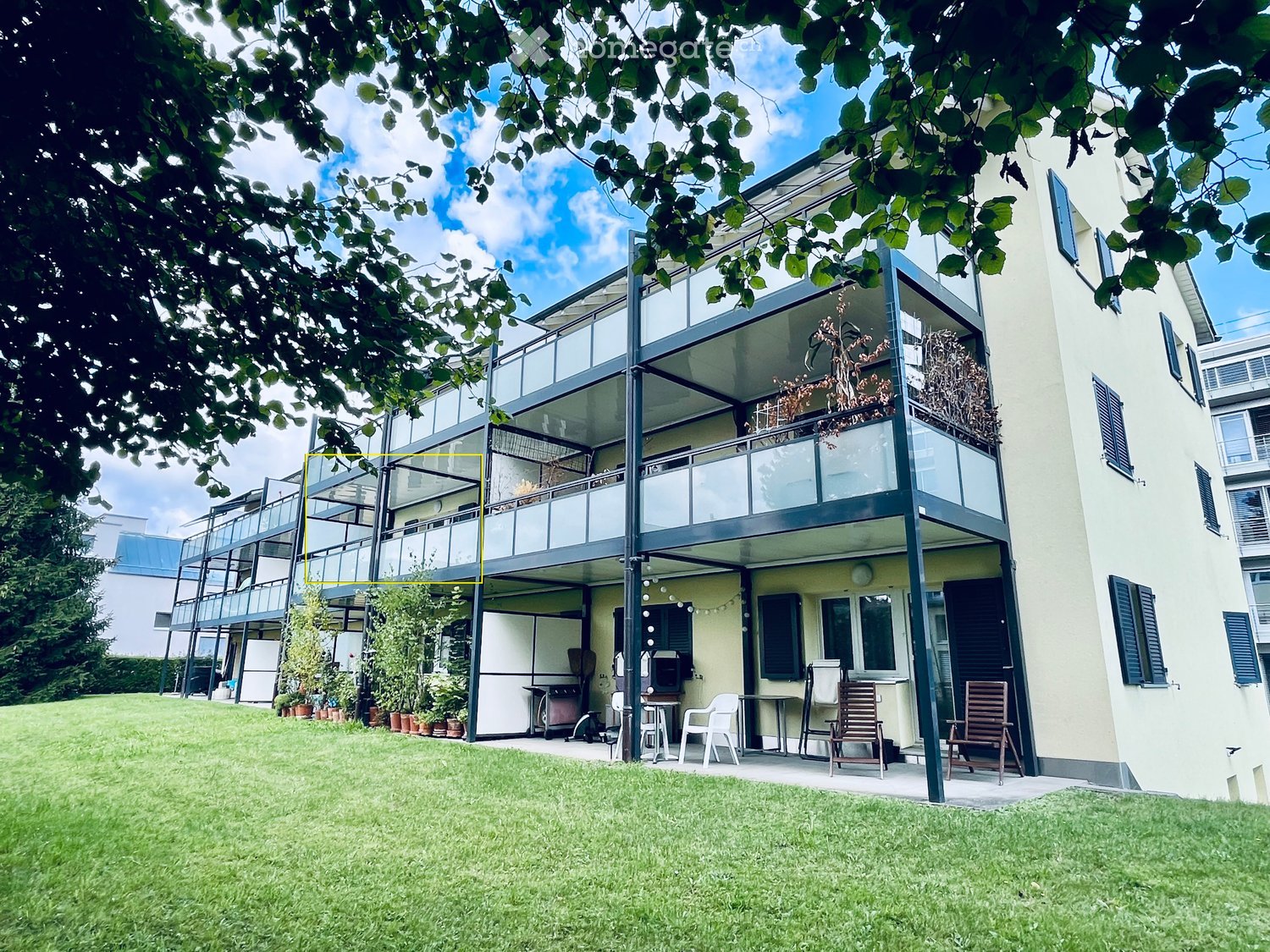 A modern two-story house with a spacious garden and outdoor seating area. The garden features a well-maintained lawn, several potted plants, and a patio with tables and chairs. The house has large windows and a balcony with glass railings.