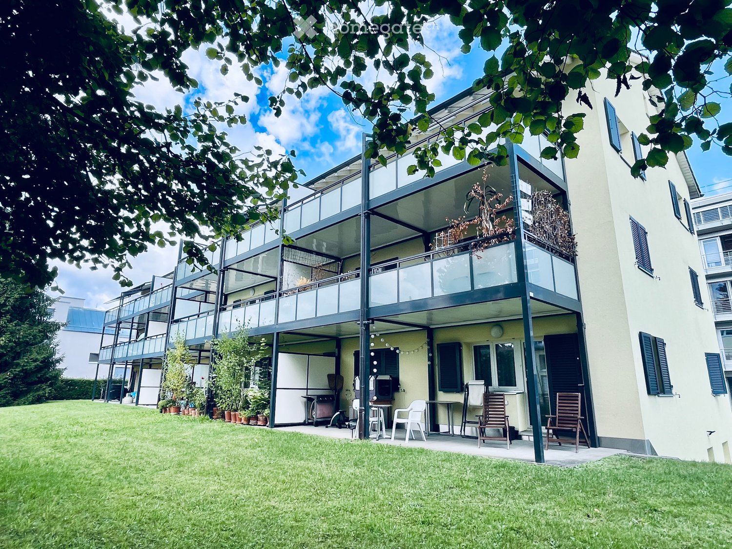 A modern building with a spacious outdoor area featuring a grassy lawn, patio furniture, and a raised balcony with glass railings.