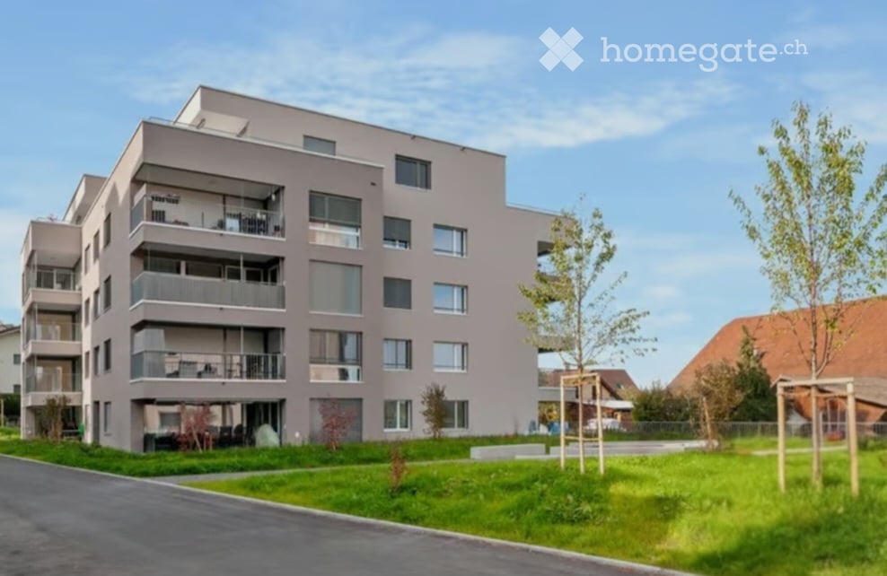 Modern multi-story apartment building with balconies, large windows, and a landscaped green area in front.