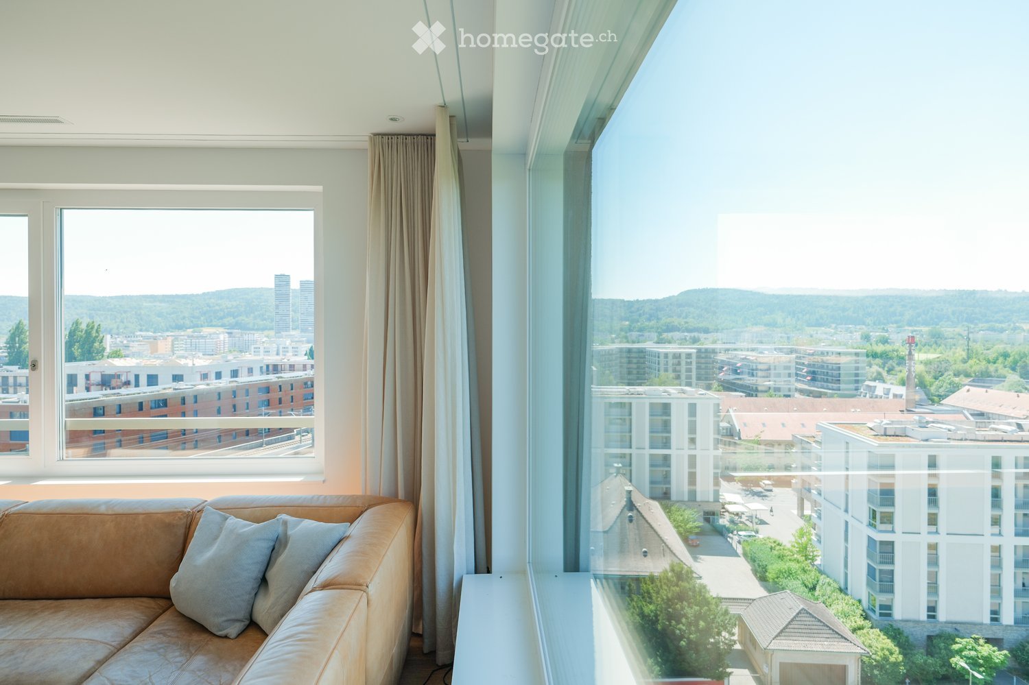Modern living room with large windows offering a panoramic city view, featuring a brown leather sofa with light-colored cushions.