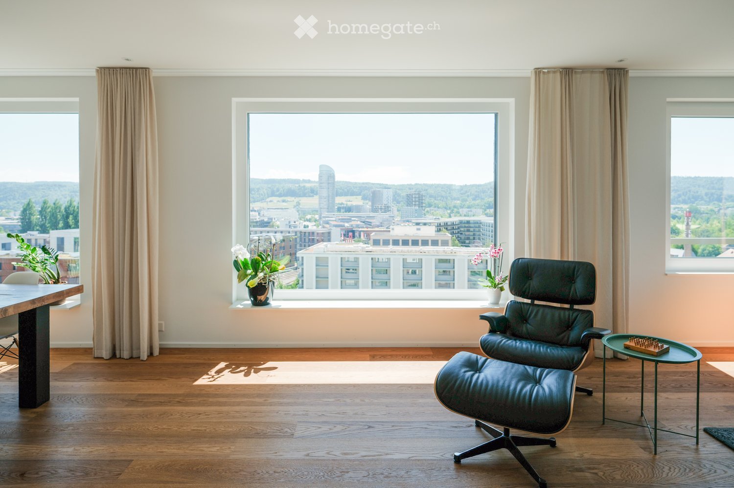 A modern living room with wooden flooring, large windows offering a city view, a black leather chair, a small coffee table, and a dining table with chairs.