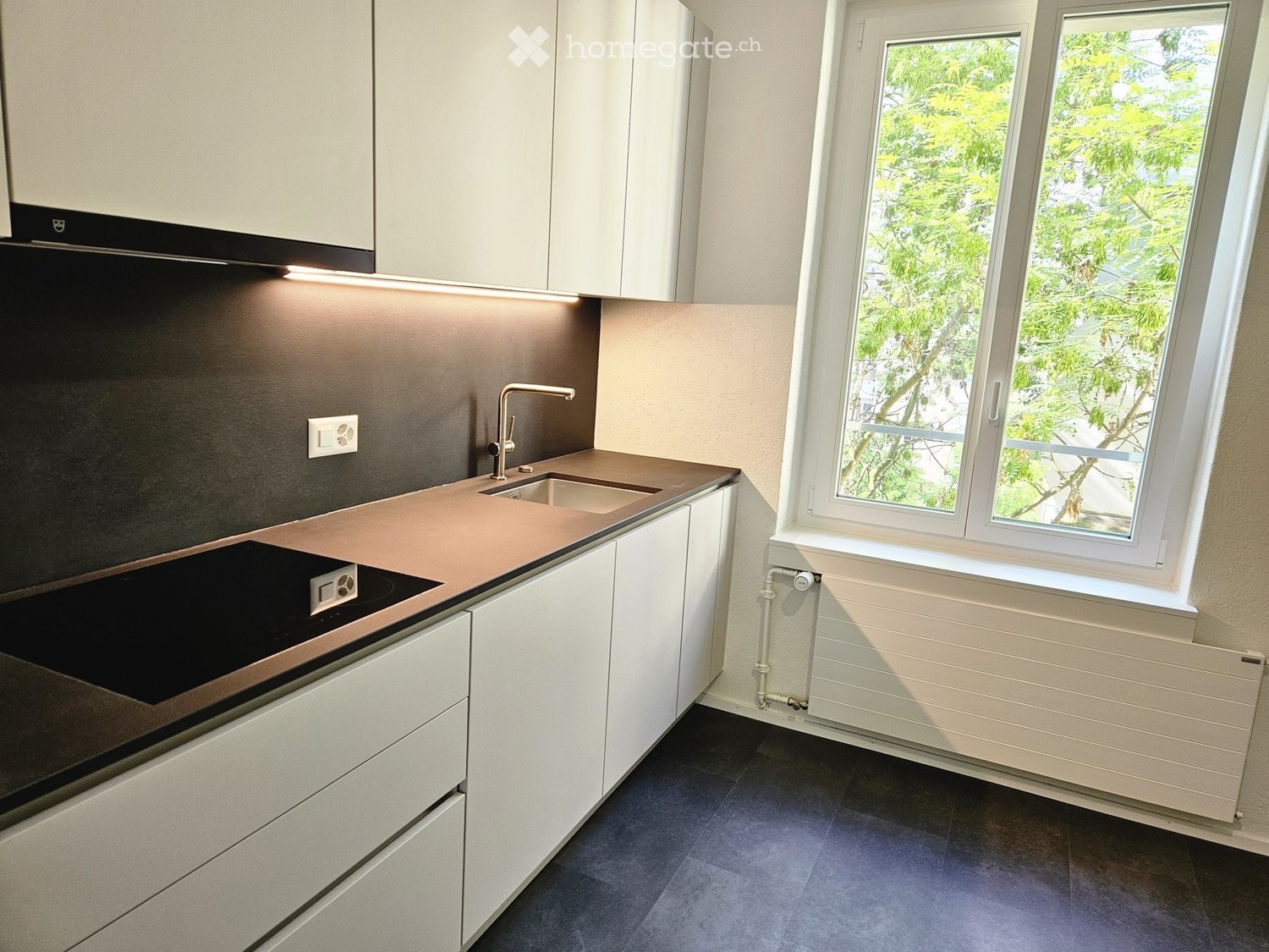 Modern kitchen with white cabinets, dark countertops, a sink, and a stove. Large window with a view of greenery.