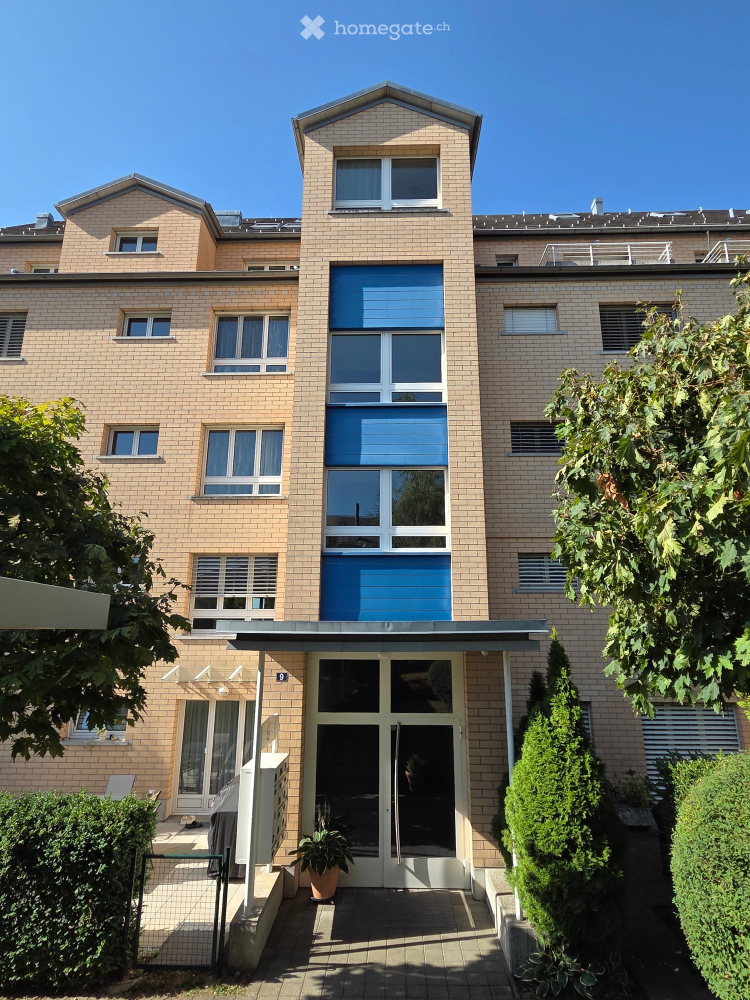A modern, multi-story brick building with large windows and a central entrance. The facade features a mix of light and dark brick patterns, with a small garden area in front.