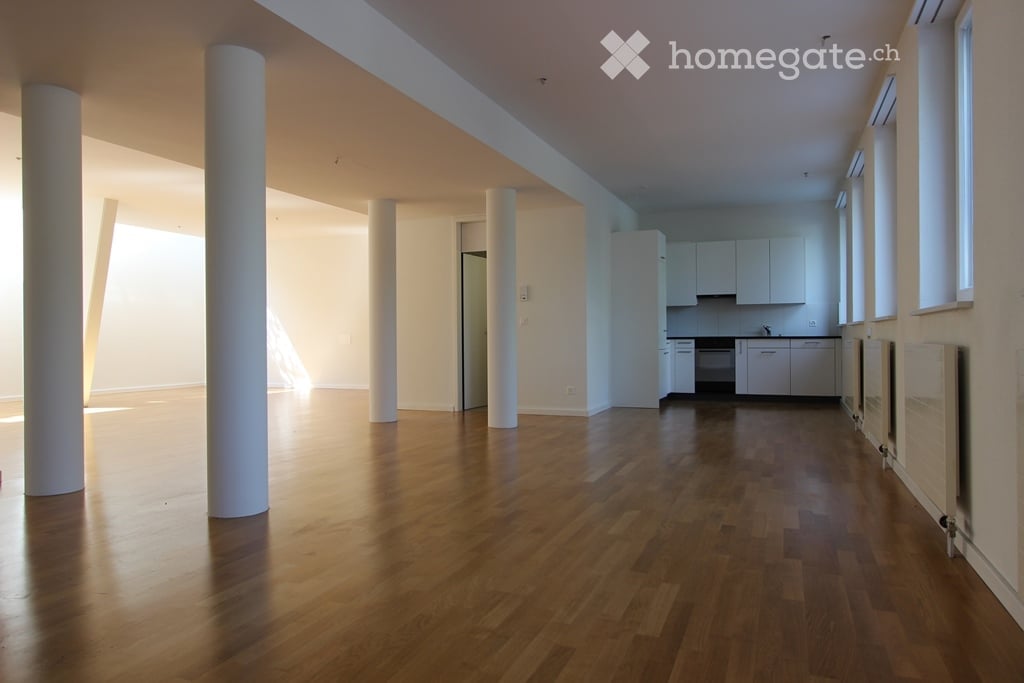 Open-plan unfurnished space with wooden flooring, white walls, and a small kitchen area featuring white cabinets and a sink.