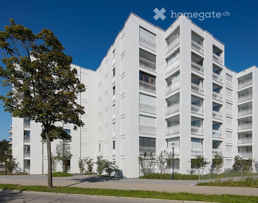 Modern white apartment building with multiple floors, balconies, and large windows. The building is surrounded by greenery and a paved area.