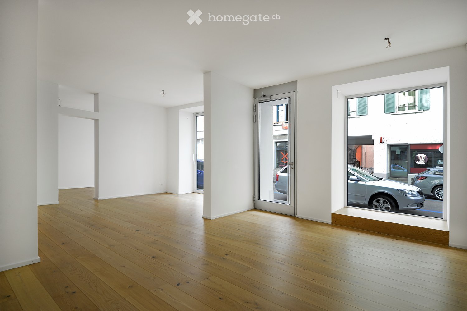 Empty room with white walls and wooden flooring, featuring large windows and a glass door leading outside.