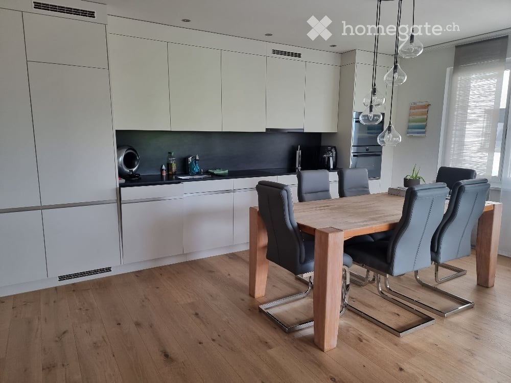 Modern kitchen and dining area with white cabinets, wooden dining table, grey chairs, pendant lights, and stainless steel appliances.