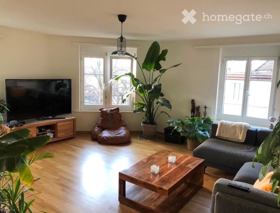 Bright living room with wooden flooring, large windows, a grey sectional sofa, a wooden coffee table, a bean bag chair, and a TV stand.
