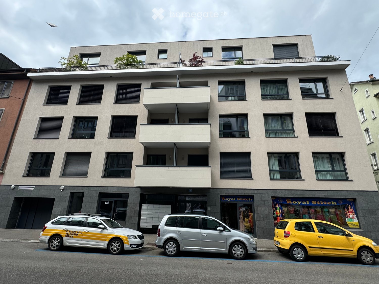 Modern multi-story building with balconies, large windows, and a rooftop terrace. Three cars are parked in front.